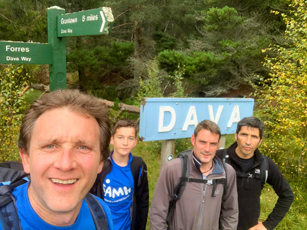Four men pose for a selfie in front of a walking route signpost