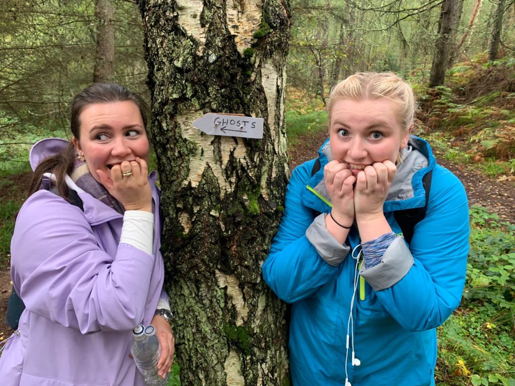 Two women stand next to a tree looking mock scared with a metal sign saying ghosts written on it