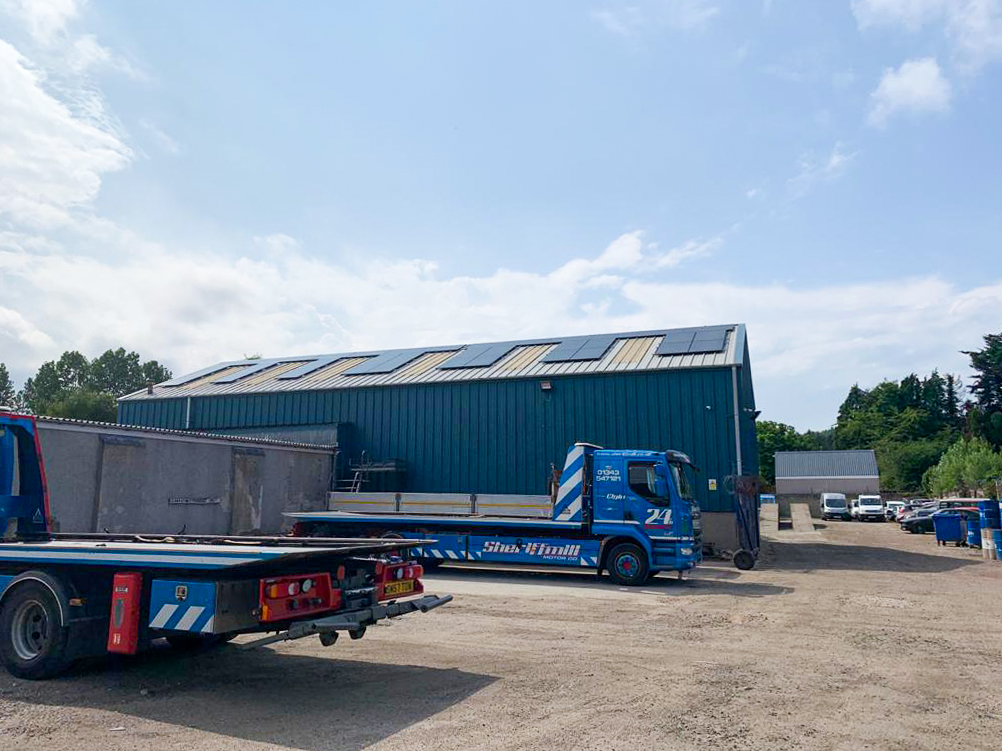 A large commercial building with solar panels on the roof and two smaller lorries parked outside.