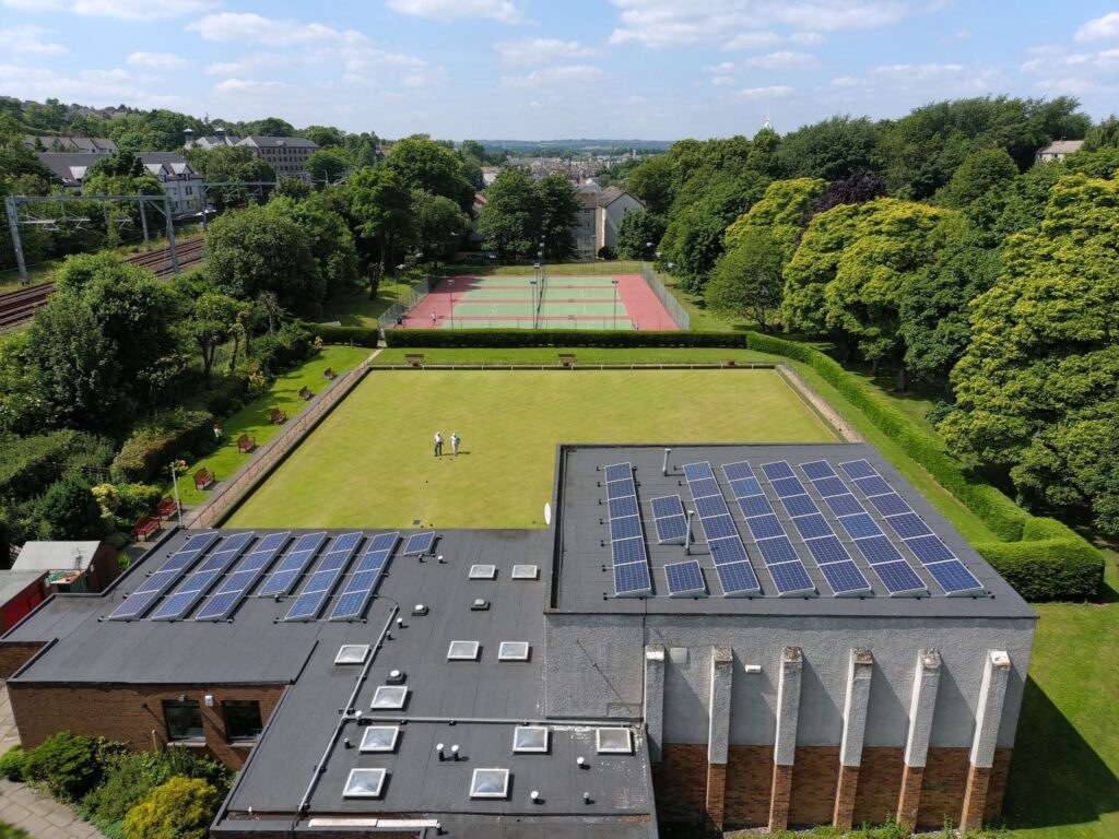 An aerial shot of a bowling green with two people walking across with tennis courts in the background. The building in the foreground is Linlithgow Sports Club and has Solar PV panels installed on the roof.