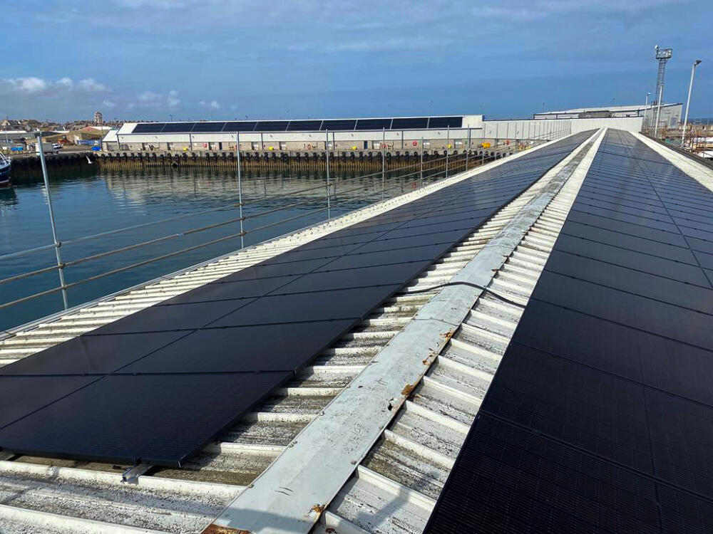 A photo taken on a large commercial roof of a building at the harbour with solar panels on the roof. In the background, you can see another solar panel system on another large building.