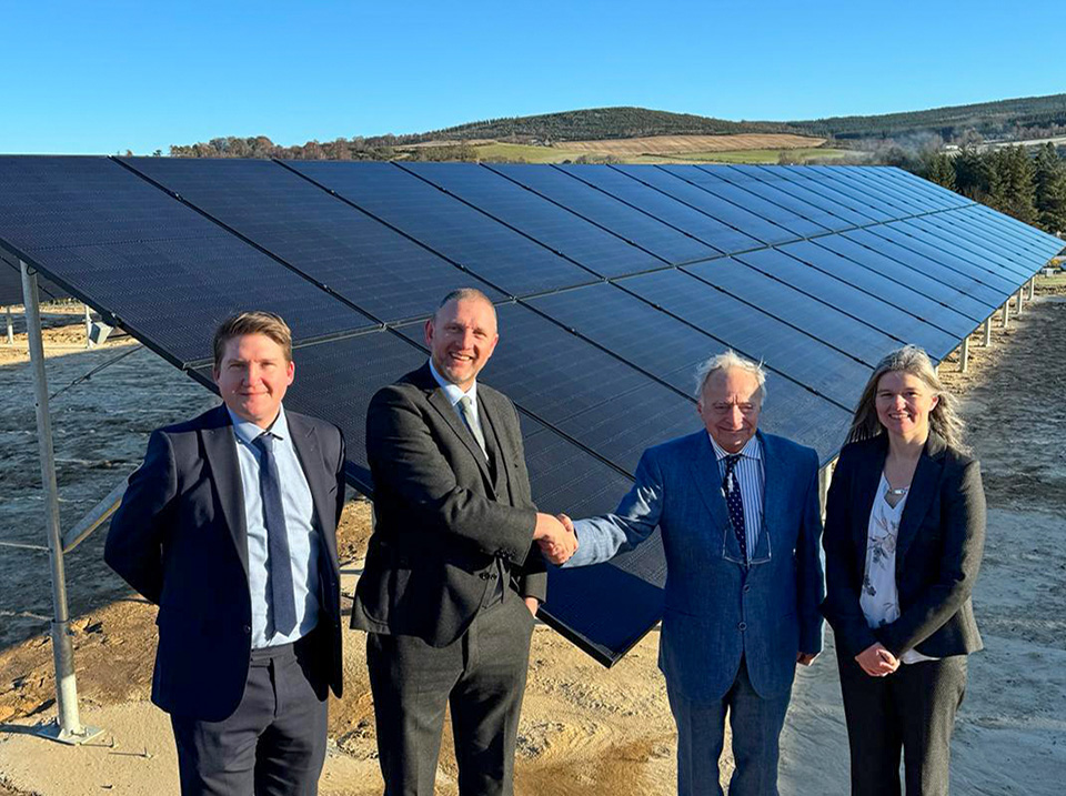 AES Solar Founder Steps Down in an image which shows four people gathered around a ground mount solar PV system with the two men in the centre shaking hands.