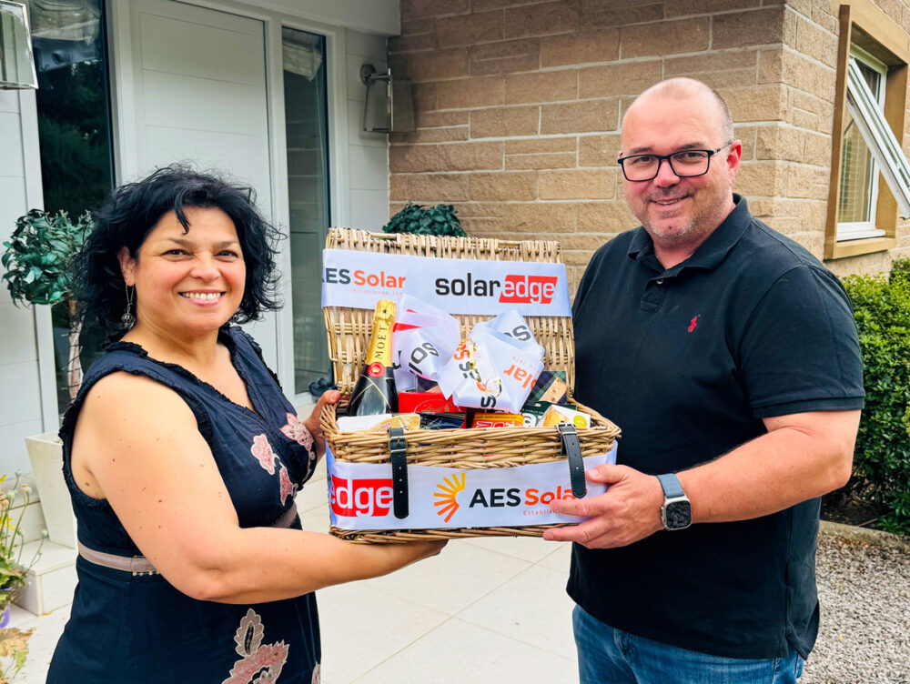 A woman and a man stand in front of a house with the woman gifting a hamper to celebrate our 1000th SolarEdge Installation
