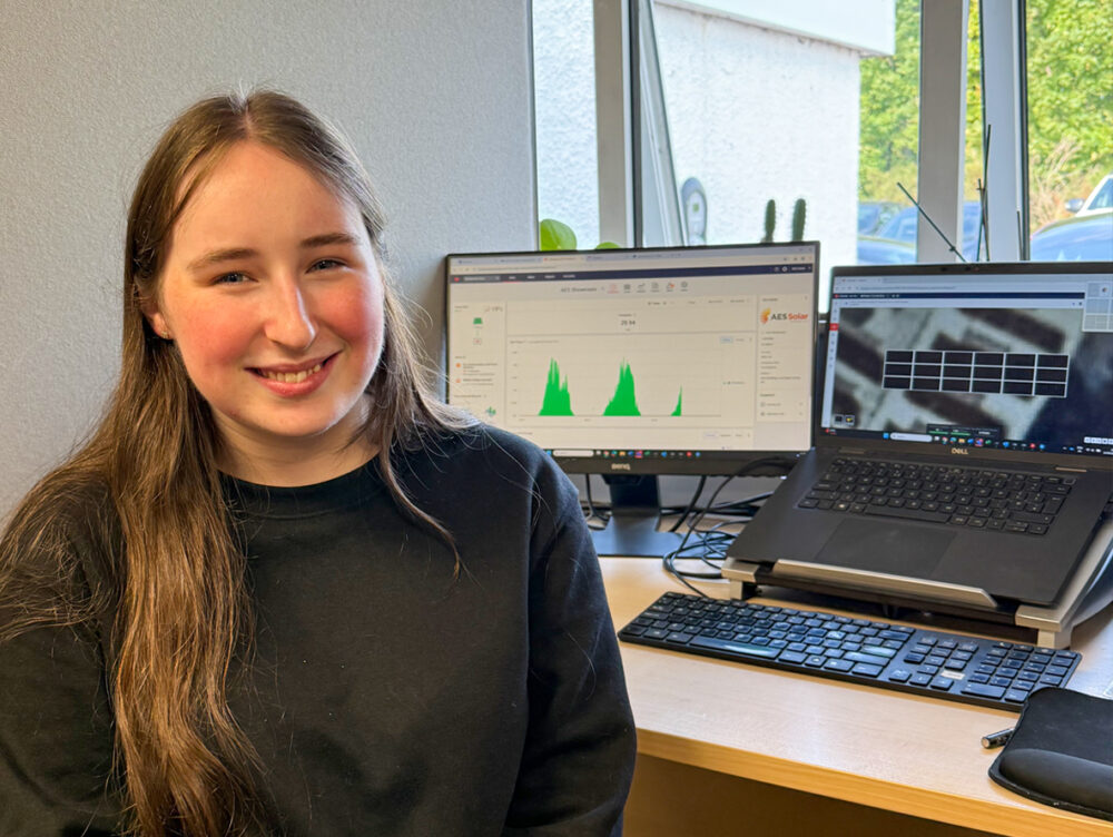 A young woman sits at a desk facing the camera on work experience at AES Solar with two computer screens in the background.