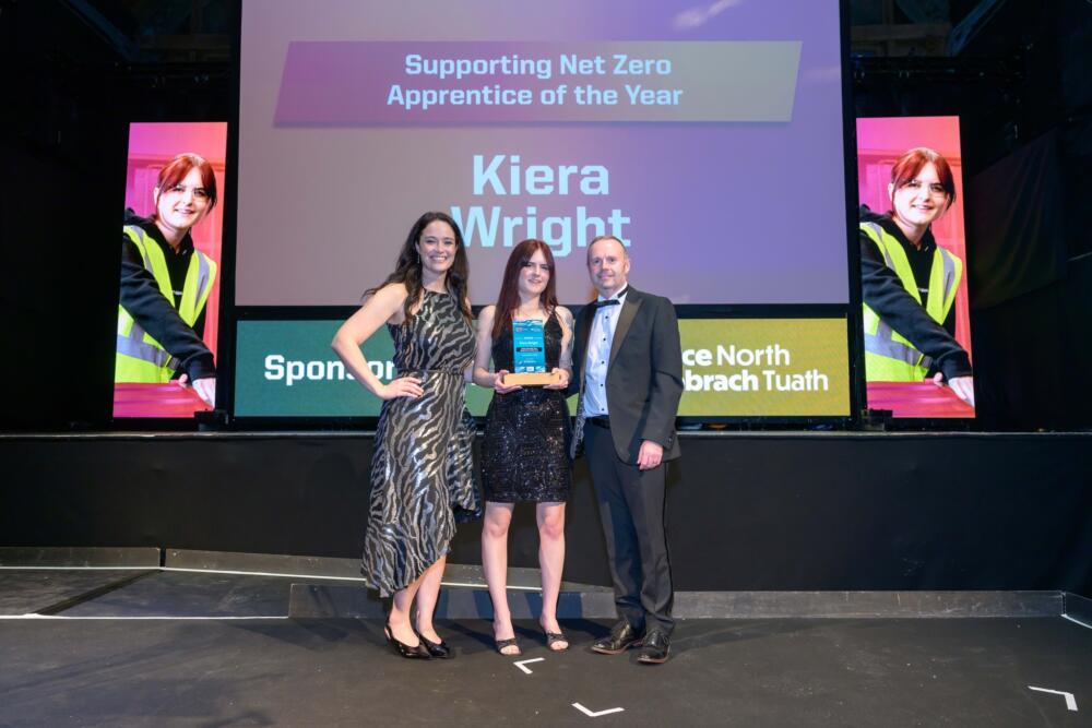 A young woman stand in the centre of another woman and a man holding a trophy recognising her efforts in inspiring the next generation.